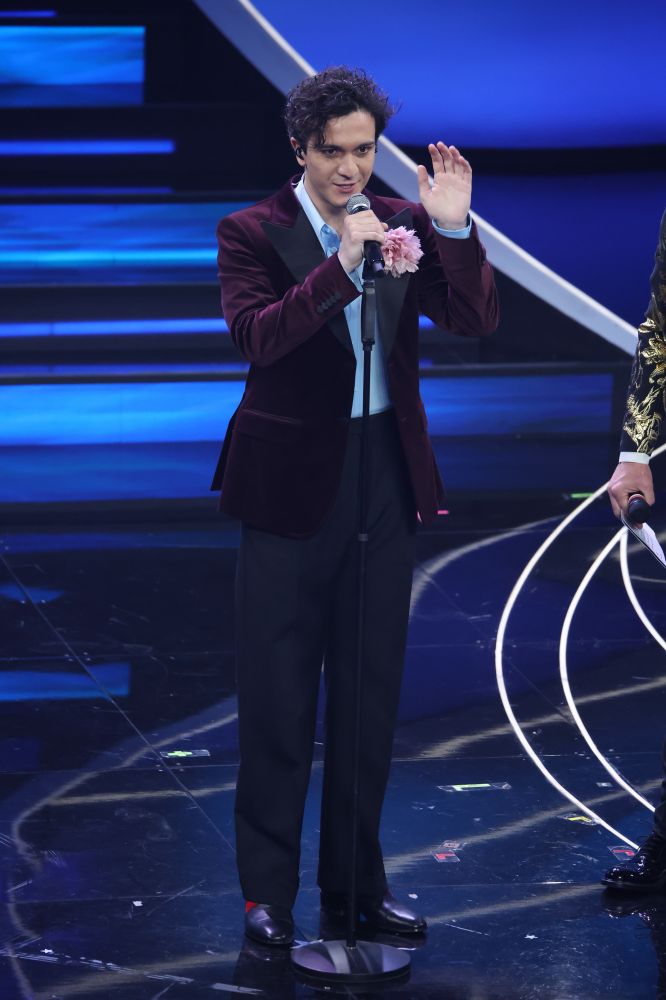 Una versione romantica del bad boy. Seconda volta sul palco di Sanremo e in gara con la ballata “Tango”, Tananai indossa un total look Gucci vestito dallo stylist Nick Cerioni. Elegante e un filo retrò la silhouette si compone con giacca con revers anni Settanta in velluto viola accessoriata con una voluminosa spilla peonia rosa sotto una camicia azzurra a contrasto e un paio di pantaloni sartoriali grigi.